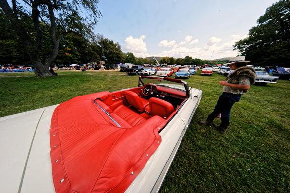 Wide angle shot from the behind a white convertable with an orange interior with a young man with a hat and racoon skin standing to the left and a few big trees and numerous classic cars on a grass field with cumulus clouds far away under a blue sky