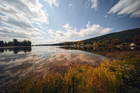 Wide angle shot of a lake with grass on the lower right,  a body of water with sky and clouds reflected in it,  a dark strip of trees coming in from the left,  a typical upstate New York hill on rising on the right with some lake houses on the shore and stretched out cumulus clouds in a blue sky above.