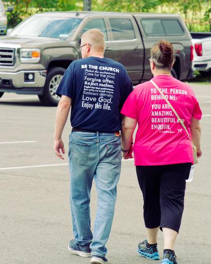 We're looking at the back of a couple holding hands in a parkihng lot with a big grey pickup truck with a covered bed behind them.  The man,  just a bit left of center is wearing jeans, sneakers and a blue shirt that reads:

BE THE CHURCH
Protect the environment.
Care for the poor.
Forgive often.
Reject racism.
Fight for the powerlessness.
Share natural and spiritual resources.
Embrace diversity.
Love God.
Enjoy this life.

To woman, to the right (his right since he's facing away) has a pink sh…