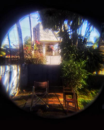 La imagen muestra una escena vista con una lente sucia a través de un objetivo de una lupa, lo que crea un efecto circular en el borde. En el centro, se observan dos sillas de madera, una de ellas plegable, situadas en un patio. A la izquierda, hay un muro que separa el patio del fondo, donde se puede ver una casa con una estructura de techo visible. Alrededor, hay vegetación, incluyendo árboles y arbustos, que añaden un toque natural al entorno. La luz del sol ilumina la escena, creando sombras y reflejos en los objetos. El cielo es visible en el fondo, con un tono azul claro. La imagen tiene un tono cálido, con predominancia de colores verdes y marrones, y presenta un efecto de distorsión característico de la visión de una lente sucia a través de un lupa.