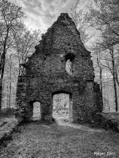 Portrait- Version: Schwarz-Weiß-Aufnahme der Kirchenruine St. Valentin bei Laubach. Die steil aufragende Frontwand mit ihren Torbögen und dem Fensterrest hebt sich eindrucksvoll vor den Bäumen und dem Himmel ab. Ein stilles Zeugnis vergangener Zeiten, das Erhabenheit und Vergänglichkeit zugleich ausst