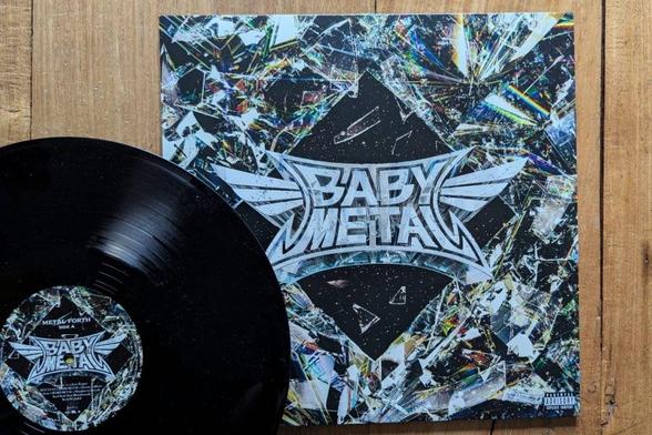 Eine schwarze Vinyl-Schallplatte neben ihrem bunten Albumcover mit einem Design aus zerbrochenem Glas und dem Text "BABYMETAL" in der Mitte.