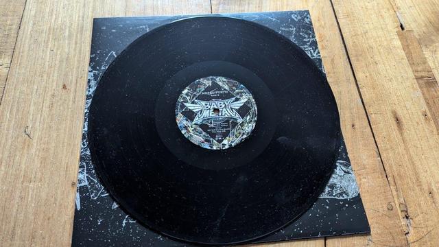 Eine Vinyl-Schallplatte, die auf einer hölzernen Oberfläche ausgestellt ist, mit einer schwarzen Scheibe und einem bunten Etikett in der Mitte. Das Etikett enthält den Namen "BABYMETAL" und den Titel "METAL FORTH". Die Schallplatte ruht teilweise auf einer verzierten Hülle.