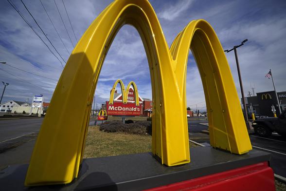 Close-up of a McDonald’s golden arches sign with another McDonald’s sign and restaurant visible in the background under a partly cloudy sky.