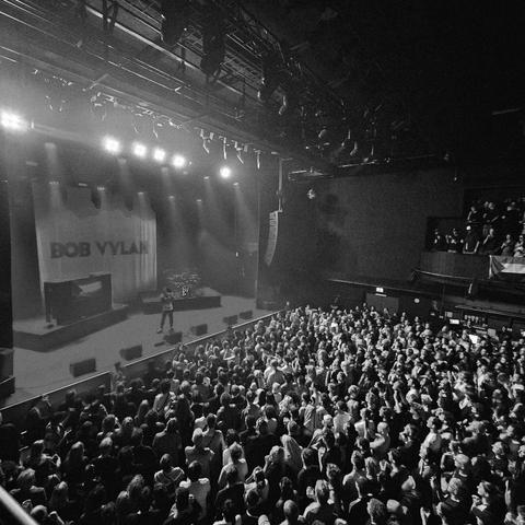 Zwart-witfoto van een concert van Bob Vylan in de grote zaal van Doornroosje. Een volle zaal met een groot publiek kijkt naar het verlichte podium, waar de artiest optreedt voor een achtergrond met de naam "Bob Vylan".