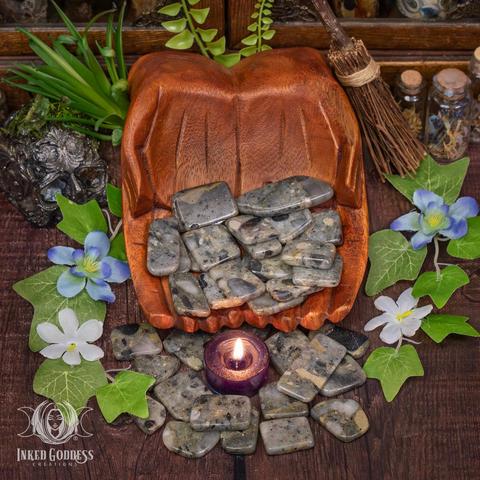 Several African Gold Pyrite slices rest in cupped, carved wooden hands, while other specimens spill onto a wooden surface. They are roughly rectangular and tumbled smooth in a wide range of greys and browns. Presented by Inked Goddess Creations.