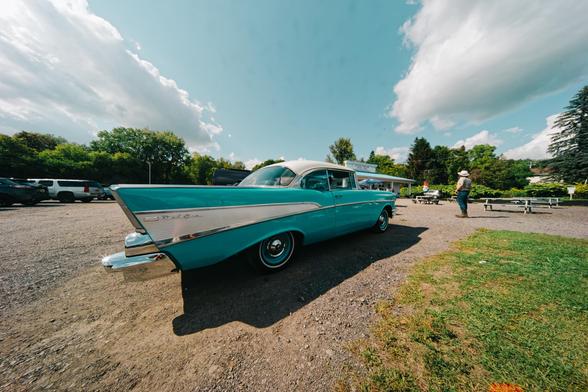 Big blue car with tailfins with a white stripe expanding from the front to to the back of the tailfin parked on a gravel parking lot,  a man in a hat standspast it next to an outdoor table and a soft serve shack is beyond that under a sky with two big cumulus clouds.