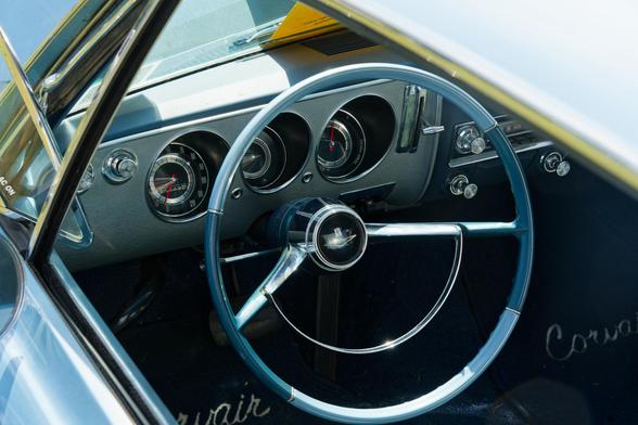 Looking into the driver's side of a car from the side window where we can see the whole circle of the streering wheel with three recessed dials beyond it and a dew circular knobs and footmats that say Corvair.