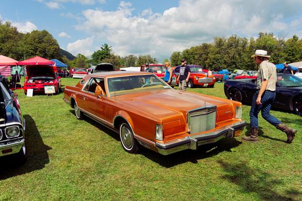 Big orange blocky car with a solid looking grille and door closed over headlights parked in the middle of a grassy field with cars on all sides of it and a man in a hat walking past it.