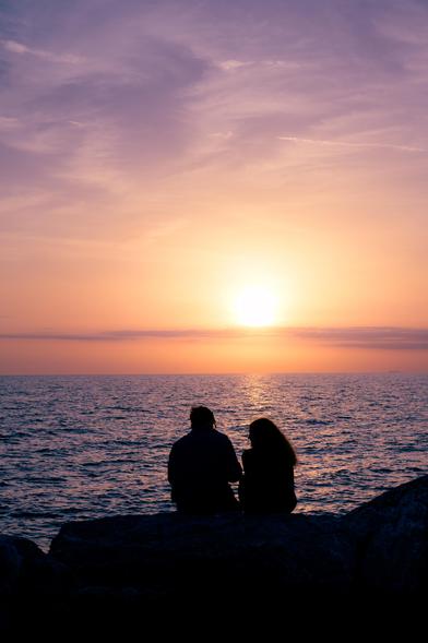 Sonnenuntergang am Ligurischen Meer in Pisa. Ein Paar sitzt auf den Steinen am Strand und schaut in den Sonnenuntergang. Es ist nur als Silhouette zu sehen. Das Wasser glitzert.