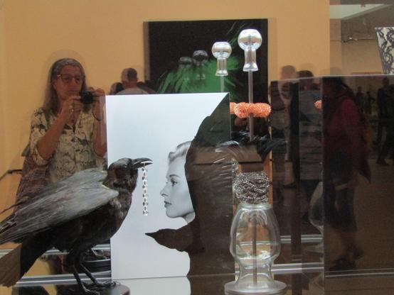 In einer Glasvitrine steht ein ausgestopfter Rabe mit Schmuck im Schnabel, ein Schwarzweiß Foto einer Dame, eine Glaskaraffe und ein anderes Foto. Im Hintergrund bin ich mit Fotoapparat zu sehen.