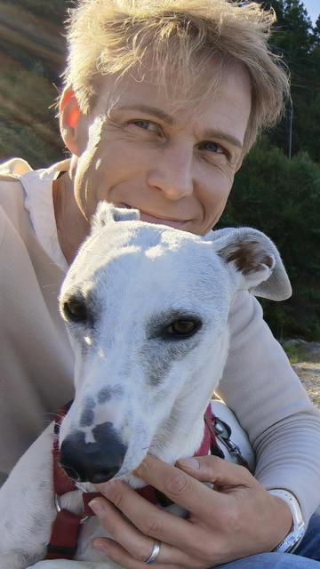 Smiling person outdoors hugging a white dog with brown spots and a red collar while the sun shines through the trees in the background