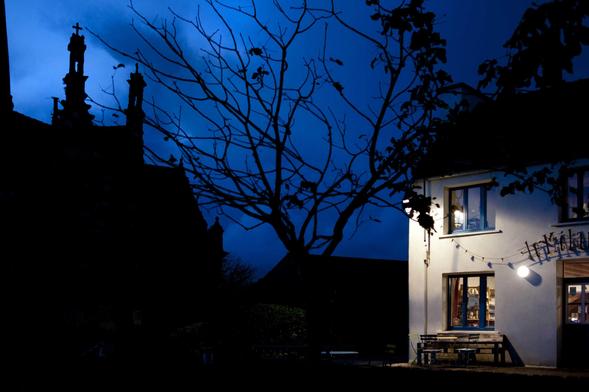 Le ciel est d’un bleu sombre, la nuit est presque tombée. Sur la gauche, se détache la silhouette noire d'une partie de l’église Saint-Mélar. On voit une croix perchée sur son pinacle, comme brandie vers le ciel. Les branches sans feuilles d’un petit arbre occupent le centre de l’image et à droite, la facade illuminée d’un bâtiment - une épicerie-café -  rassure dans cette inquiétante ambiance. 
