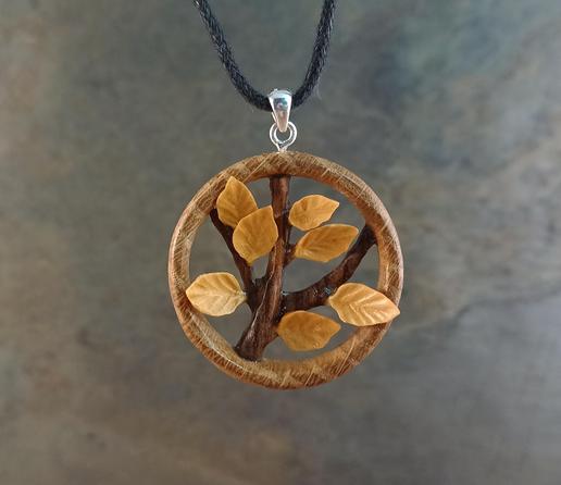 A carved wooden pendant, consisting of a small circle of light oak, with a three pronged branch of darker walnut inside it, with seven golden boxwood leaves coming from the branch. Held suspended from a black cord with a grey skate background.