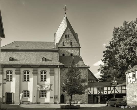 Die Evangelische Stadtkirche in Laubach erhebt sich mit ihrem wuchtigen Turm und den barocken Fenstern über den Platz der Altstadt. Im Vordergrund ein Baum und Fachwerkdetails, die der Szene eine einladende, historische Atmosphäre verleihen. Das Schwarz-Weiß des Fotos unterstreicht die Zeitlosigkeit und den Charakter des Bauwerks.