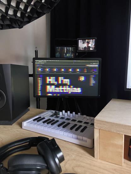 A wooden desk with headphones, a speaker, and an Arturia Minilab 3 in the front. In the back, a 20" monitor, with the Elgato Prompter directly above, as well as another smaller monitor that shows the current camera image.