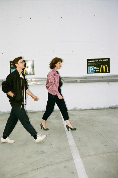 Tim Roth und Nina Schellhase im Profil. Sie laufen in einem zügigen Schritt über einen Parkplatz. An der weißen Wand hängt ein kleines Schild, das zeigt, dass es sich um einen McDonald's Parkplatz handelt. Das Foto wurde von Sebastian Schramm aufgenommen.