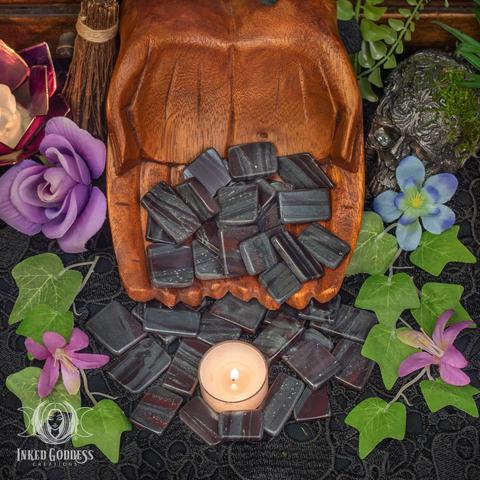 Many specimens of the "Ancestralite Slice" product are spilling from carved wooden cupped hands. They are surrounded by faux greenery and a tealight candle. They are in flat, polished, roughly rectangular shapes. They are a mix of red jasper and grey or black hematite, giving them a striped appearance. Presented by Inked Goddess Creations.