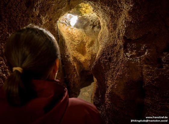 In this image, we see a person standing in a dimly lit cave, their gaze fixed on a bright light source in the distance. The cave walls are dark, creating a stark contrast with the person's figure. The dominant colors in the scene are black and brown, with the accent color being a deep shade of brown. The person, identified as the main object in the image, occupies a significant portion of the frame and appears to be captivated by the mysterious light. The setting suggests a sense of exploration…