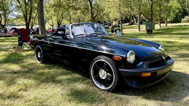 Photograph of a black 1976 MGB facing right in a park with green trees and grass.