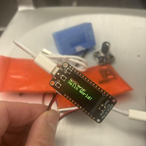 A hand holds a small electronic device displaying "Hello World!" on its screen. Surrounding it are a blue component, red plastic, a white cable, and other electronic parts.