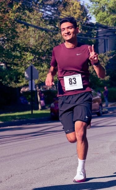 South Asian man wearing a purple Nike shirt,  a white bib with the number 83 in black and dark shorts is running on a downtwon street with two fingers of his left hand extended (your right)