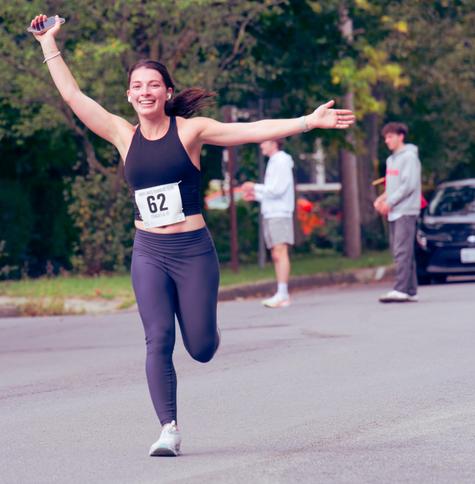 Woman wearing a black halter top with a bit of an open midriff and something like like Yoga pants has her left (your right) arm out horizontally with her palm open and her other hand up diagonally with a bracelet and holding up a phone,  she's smiling and has a ponytail suspended behind her,  her left leg is up and her right down straight down.  Behind her are two of the race volunteers controlling traffic.