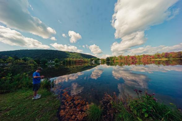 The horizon is right on the midline of this photo but a hill looms over it on the left half and above that a mostly clear blue sky with a few big clouds which are reflected on the still surface of a pond,  right near the horizon can be found a row of trees that are just starting to turn color,  on the left is the side of the pond and a boy with a blue shirt looks really tiny compared to the landscape as he fishes.