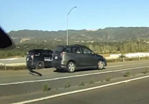 A dark-colored Toyota Matrix drives on a highway with a police SUV following behind, set against a backdrop of hills and clear skies.