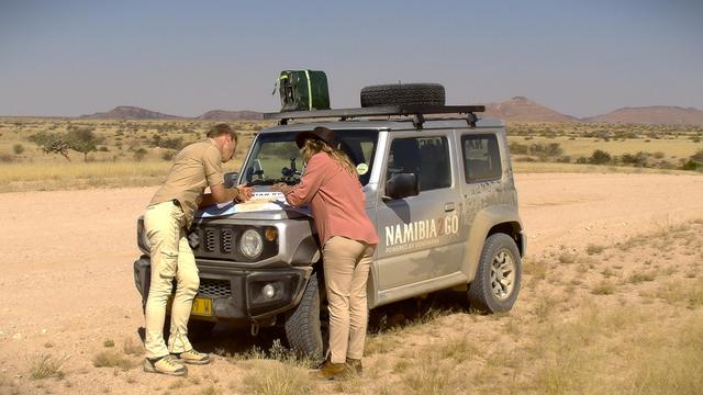 Two people in safari clothing look at a map on the hood of an off-road vehicle with the inscription “Namibia2Go” in a dry savanna landscape with a wide horizon and scattered bushes.