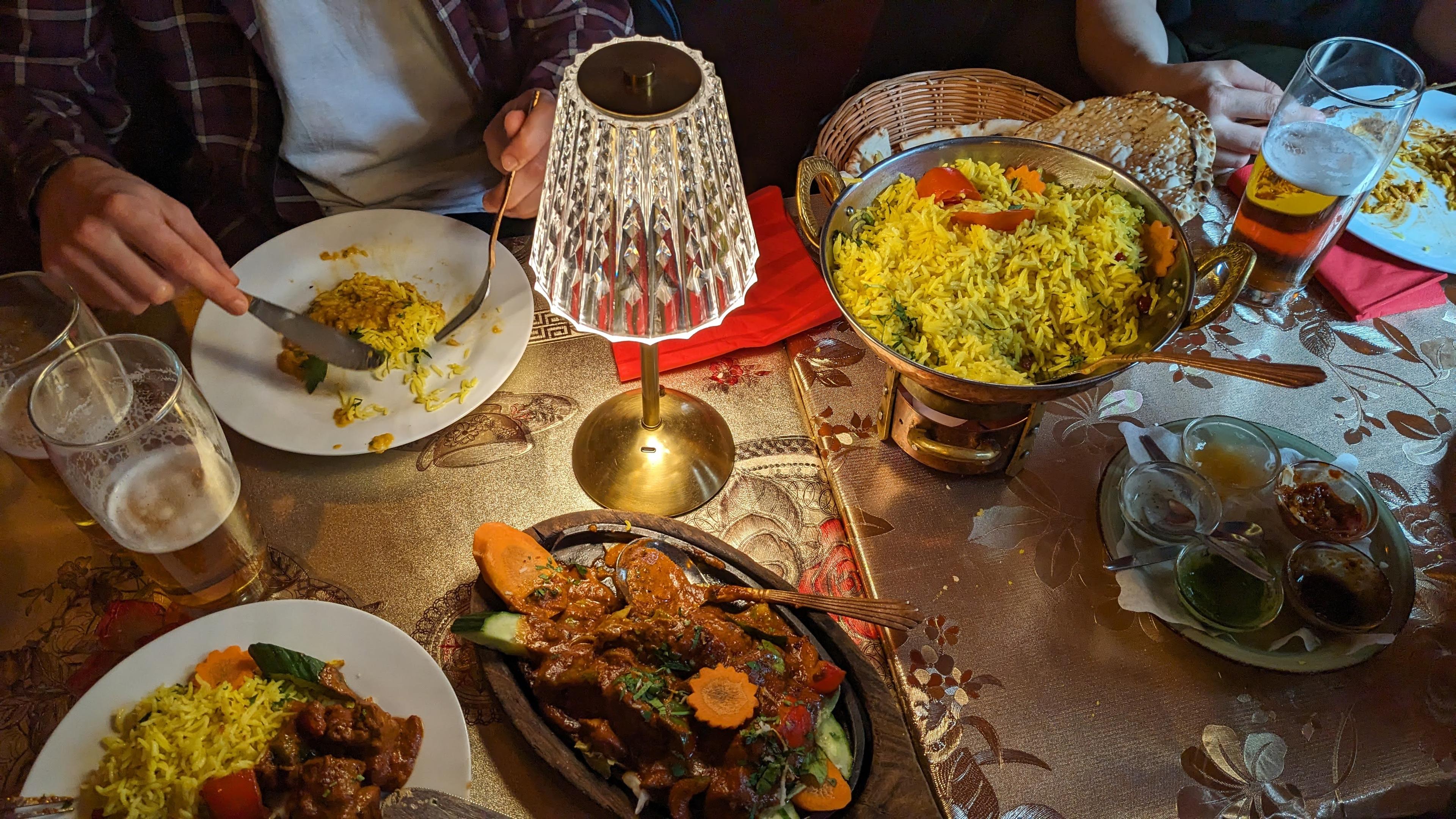 Bildbeschreibung von KI:
Das Foto zeigt eine gedeckte Restauranttischszene mit indischem Essen. In der Mitte des Tisches steht eine goldfarbene Schale mit zwei Henkeln, die randvoll mit gelbem Reis gefüllt ist, dekoriert mit Tomatenstücken und Möhrenscheiben. Daneben liegt ein Korb mit Fladenbrot. Vorne links im Bild sieht man einen Teller mit Reis und einem Fleischgericht in brauner Soße, garniert mit Möhren- und Gurkenscheiben. Rechts unten steht ein Teller mit fünf kleinen Schälchen verschiedener Soßen und Dips in unterschiedlichen Farben, darunter grün, rot und braun. In der Bildmitte leuchtet eine Tischlampe mit goldenem Fuß und einem gläsernen Schirm, die warmes Licht verbreitet. Links sitzt eine Person im karierten Hemd, die mit Gabel und Messer isst, vor ihr stehen zwei große Biergläser, teilweise geleert. Rechts sitzt eine weitere Person, deren Arme zu sehen sind, vor ihr ein Glas mit hellem Bier und ein Teller mit Essensresten. Die Tischdecke ist golden mit floralen Mustern. Die Atmosphäre wirkt gemütlich, gesellig und nach einem Abendessen im Restaurant.
Bildbeschreibung von KI:
Das Foto zeigt eine gedeckte Restauranttischszene mit indischem Essen. In der Mitte des Tisches steht eine goldfarbene Schale mit zwei Henkeln, die randvoll mit gelbem Reis gefüllt ist, dekoriert mit Tomatenstücken und Möhrenscheiben. Daneben liegt ein Korb mit Fladenbrot. Vorne links im Bild sieht man einen Teller mit Reis und einem Fleischgericht in brauner Soße, garniert mit Möhren- und Gurkenscheiben. Rechts unten steht ein Teller mit fünf kleinen Schälchen verschiedener Soßen und Dips in unterschiedlichen Farben, darunter grün, rot und braun. In der Bildmitte leuchtet eine Tischlampe mit goldenem Fuß und einem gläsernen Schirm, die warmes Licht verbreitet. Links sitzt eine Person im karierten Hemd, die mit Gabel und Messer isst, vor ihr stehen zwei große Biergläser, teilweise geleert. Rechts sitzt eine weitere Person, deren Arme zu sehen sind, vor ihr ein Glas mit hellem Bier und ein Teller mit Essensresten. Die Tischdecke ist golden mit floralen Mustern. Die Atmosphäre wirkt gemütlich, gesellig und nach einem Abendessen im Restaurant.