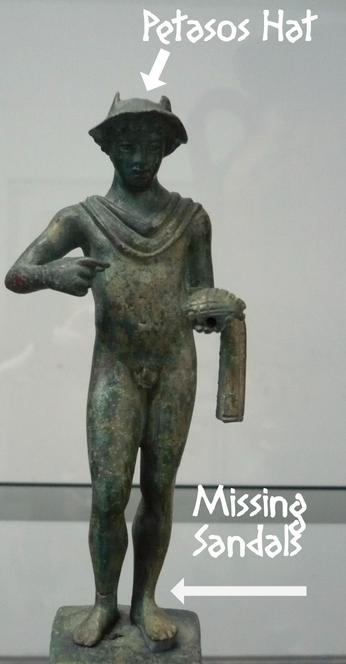 Bronze figurine of Hermes with a green patina. He is depicted in the nude, wearing just a hat and a chlamys cloak around his shoulders and draped around his left arm. He holds a purse in is left and probably used to hold a kerykeion staff in his right that is now missing.
I added the words "Petasos Hat" with an arrow to the hat and "Missing Sandals" with an arrow pointing to his naked feet in white letters.