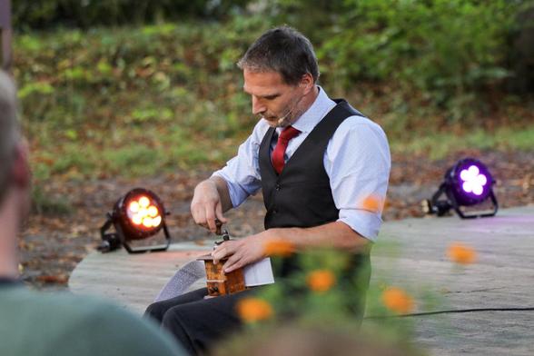 Thomas, mit hellblauen Hemd, roter Krawatte und schwarzer Weste, sitzt am Bühnenrand und kurbelt eine Kaffeemühle. Beim genaueren hinsehen erkennt man, dass es sich um eine Lochbandspieluhr handelt.