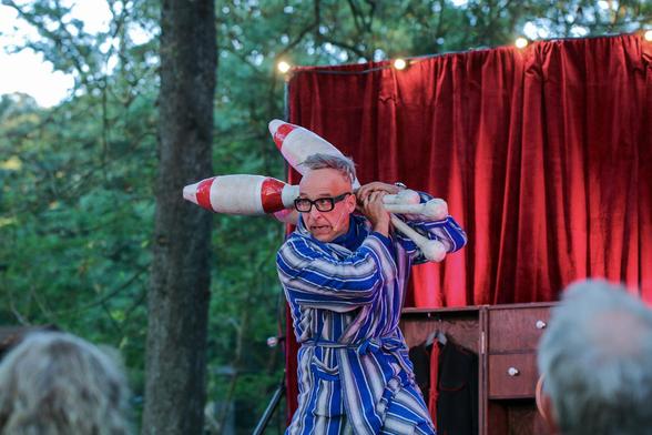 Arthur, mit schwarz gerahmter Brille und blau/weiß gestreiften Bademantel, post mit drei Riesenkeulen, die er jonglieren wird.