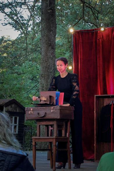 Sia, ganz in schwarz gekleidet, bedient ein Pop-Up Buch. Das Buch liegt auf einem alten Koffer. Der Koffer liegt auf einem Hocker. Der Hocker steht auf einem Stuhl. Ein improvisierter Tisch. Im Hintergrund sieht man Bäume und einen roten Vorhang, an dessen Kanten warmweiße Glühlampen leuchten.