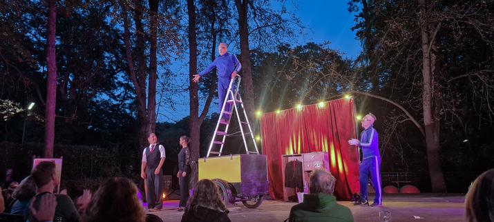 Die Abschlußszene: In der Mitte steht ein altes Lastenrad. Auf dem Koffer des Lastenrades steht eine Stehleiter. Auf der Stehleiter steht Arthur in einem blauen Trainingsanzug. Links neben dem Lastenrad stehen Sia & Thomas. Auf der anderen Seite ein Zuschauer, der sich als Arthur verkleidet hat. Im Hintergrund sind Bäume. Es ist schon dunkel. Alle stehen vor dem roten Vorhang und nehmen Applaus entgegen. Die Szene ist gut ausgeleuchtet.