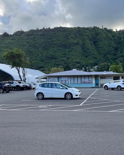 A white 2010 Honda Fit Sport edition car parked in an empty lot with mountainous greenery in the background. There are several other vehicles in the lot, and a building with a registration sign is visible nearby. The sky is partly cloudy.