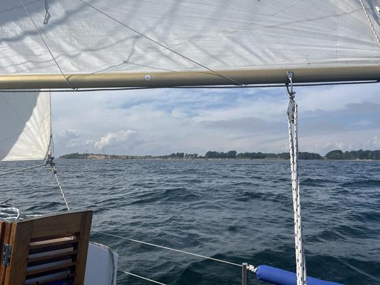 Die Flensburger Außenförde bei Broager, von einem kleinen Segelboot aus gesehen. Im Hintergrund Land, davor das Meer, der obere Rand des Bildes zeigt das Großsegel des Bootes.