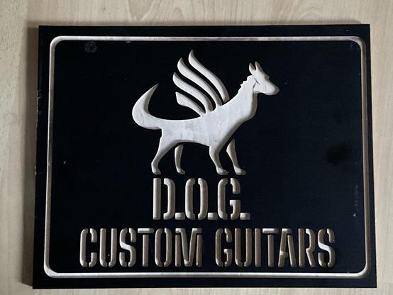 Ein dunkelbraunes Holzschild, in das das Logo von D.O.G. Custom Guitars, ein geflügelter Hund, und die Worte D.O.G. Custom Guitars gefräst sind, so dass die Schrift hell erscheint.