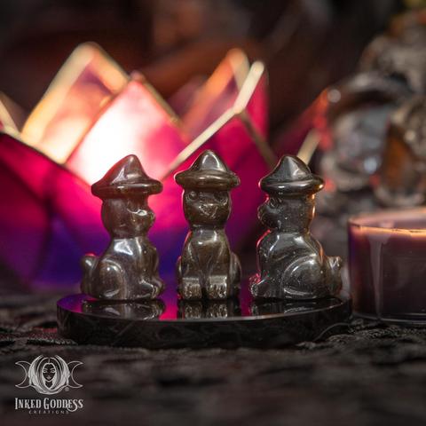 Three specimens of the "Obsidian Mini Cat with Witch Hat" product are arranged on an onyx disc. They are very small and polished and carved into the shape of a sitting cat wearing a pointed witch hat. Their color ranges from dark brown or bronze to black. Presented by Inked Goddess Creations.