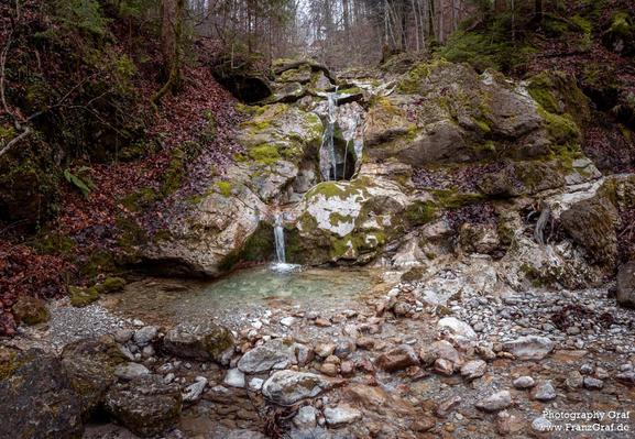 A serene and picturesque scene of a small waterfall cascading down in a rocky area is captured in this image. The rocks and boulders create a rugged yet beautiful landscape, with moss and plants adding a touch of greenery to the surroundings. The water flows gently, creating a soothing sound that adds to the peaceful ambiance. The trees in the background, along with the mountainous terrain, create a natural and untouched setting. This image showcases the beauty of nature, with the contrast of t…