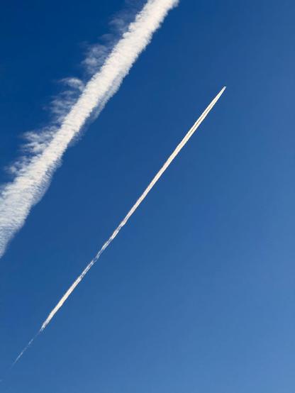 Two plane contrails are seen against a bright blue sky. The one closest to the top left of the image is older and thus wider and fuzzier. The second one is a much thinner line, and you can just make out the plane at the front of it. 