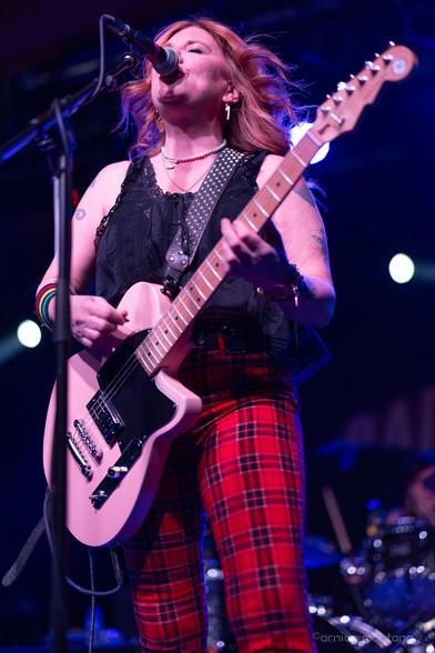 A female musician performs on stage, holding a pink guitar while singing into a microphone. She is dressed in a black top and red plaid pants, with visible accessories and tattoos. The background features colorful stage lighting.