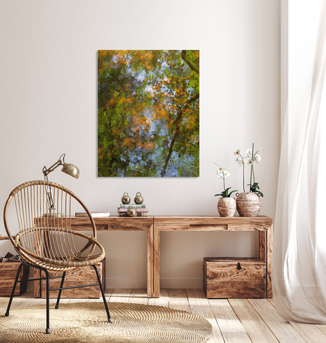 An impressionistic photograph of a pond with green and orange Autumn leaves and blue water on a white wall in a room with a wooden table and a wicker chair.