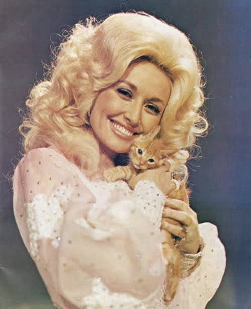 An older photo of Dolly Parton holding an orange kitten to her cheek. Dolly is smiling and wearing a white, billowy garment. Her hair is high and reaching up to God