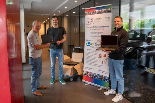 In einem Bürogebäude stehen 3 Männer vor einem Rollup von Labdoo. Jeder hält eine Laptopspende in der Hand. Auf einem Rollwagen steht ein Pappkarton mit weiteren Laptops.