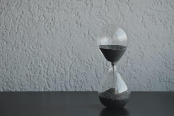 A glass hourglass with black sand running from the upper to the lower bulb, placed on a dark surface against a light textured wall.