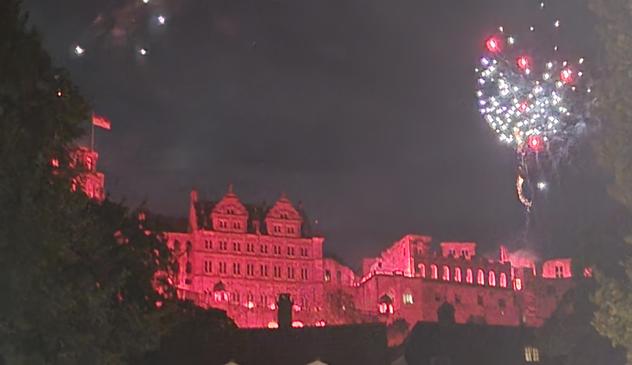 Castle overlooking the town of Heidelberg, glowing red to symbolise fire. Above are display of fireworks.  