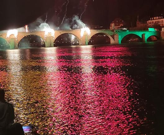 Old bridge in Heidelberg town lit by fireworks with the river Neckar appearing orange and crimson with the reflection.