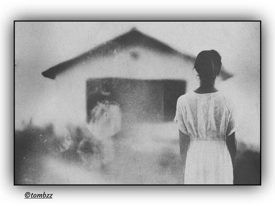 A black-and-white image with a deliberately damaged texture, achieved through soft focus and visible grain, depicts a hazy, dreamlike scene. In the foreground, a solitary figure with her back to the viewer stands wearing a loose, light-colored dress and her hair in a bun.
​The scene has a melancholic, ethereal mood. The woman gazes toward a blurry, simplified building, whose shape is barely visible in the background. To her left, in the deep shadow, the faint outline of a second, undefined figure appears. The entire composition is infused with a sense of mystery, and the scratches on the film add a raw, poignant character to the photograph.